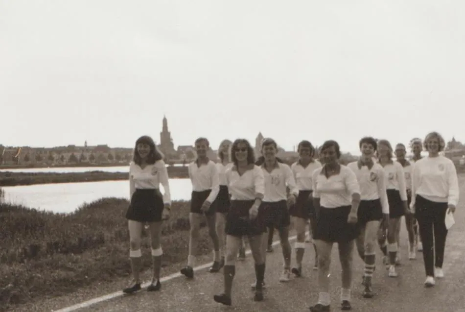 Speelsters in clubtenue lopen in een rij over een dijk, met de torens van Kampen op de achtergrond