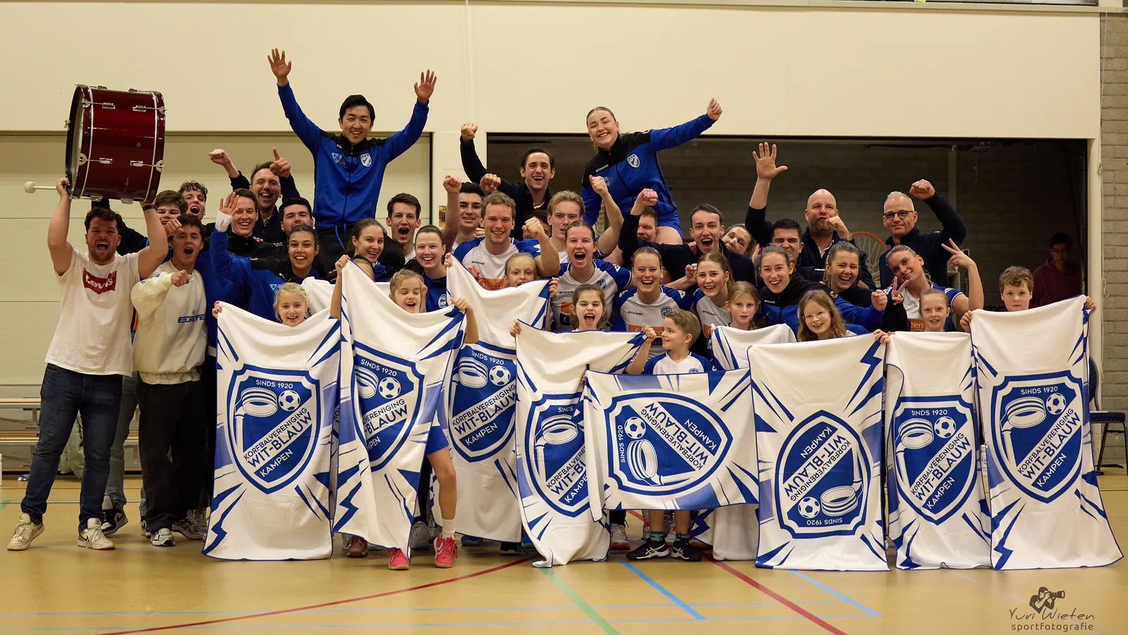 Jong en oud van Wit-Blauw juichen samen in de sporthal, met vlaggen van de club